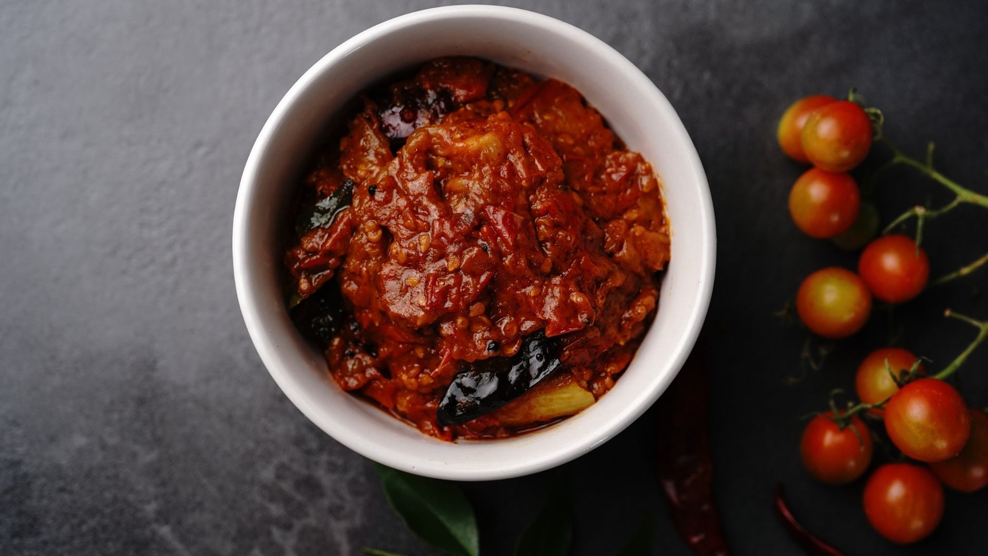 A bowl of rich, red tomato chutney with visible chili peppers and fresh tomatoes on the side.