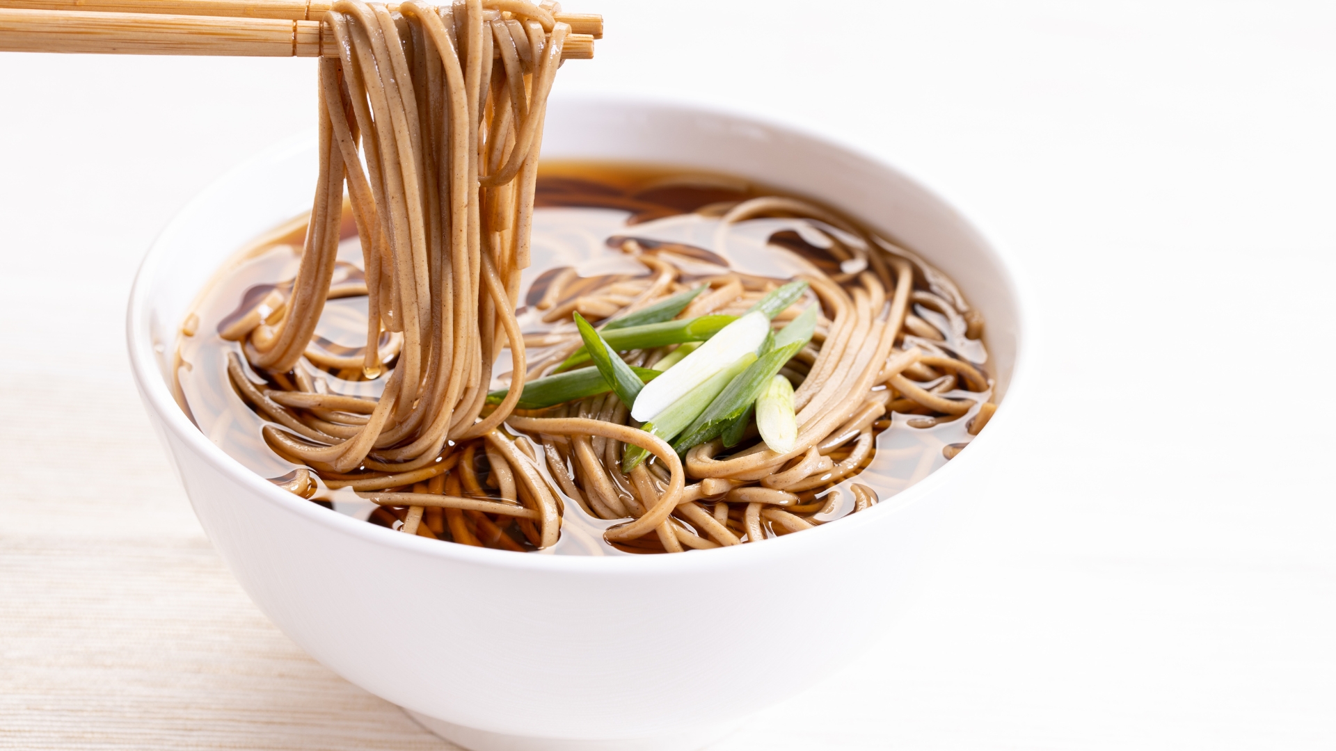  A bowl of soba noodles served in a warm broth, garnished with scallions, being picked up with chopsticks.