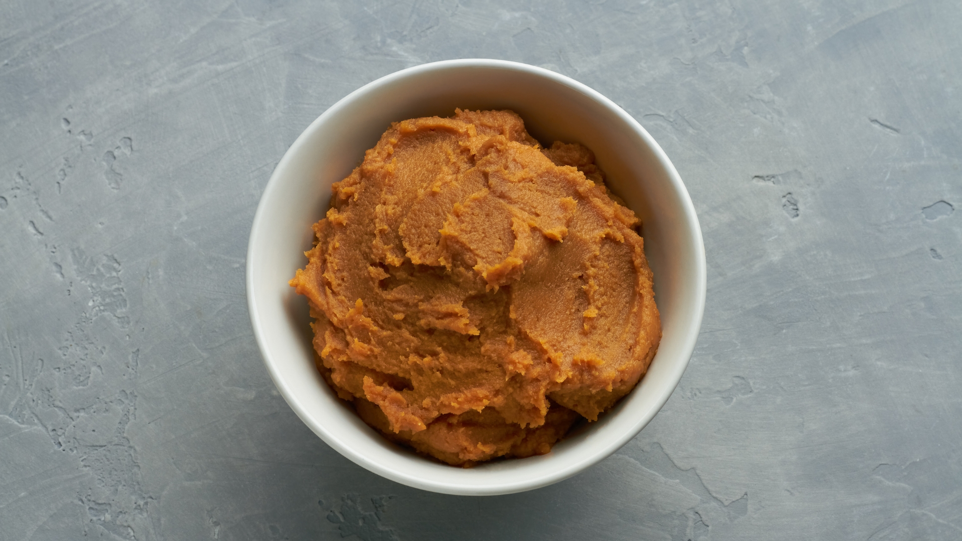 A bowl of thick, golden-brown miso paste on a gray surface, ready for use in soups and marinades.