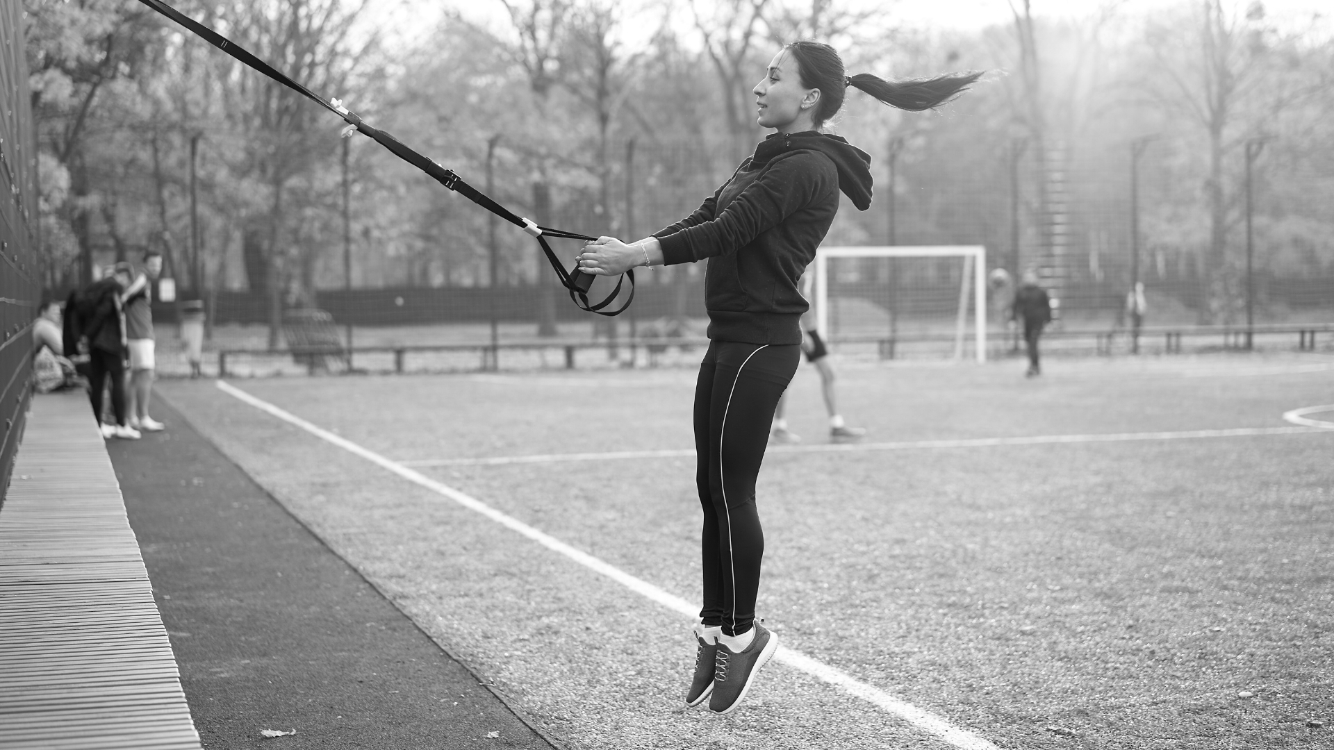 A female athlete performing a TRX-assisted jump squat outdoors, demonstrating explosive movement.