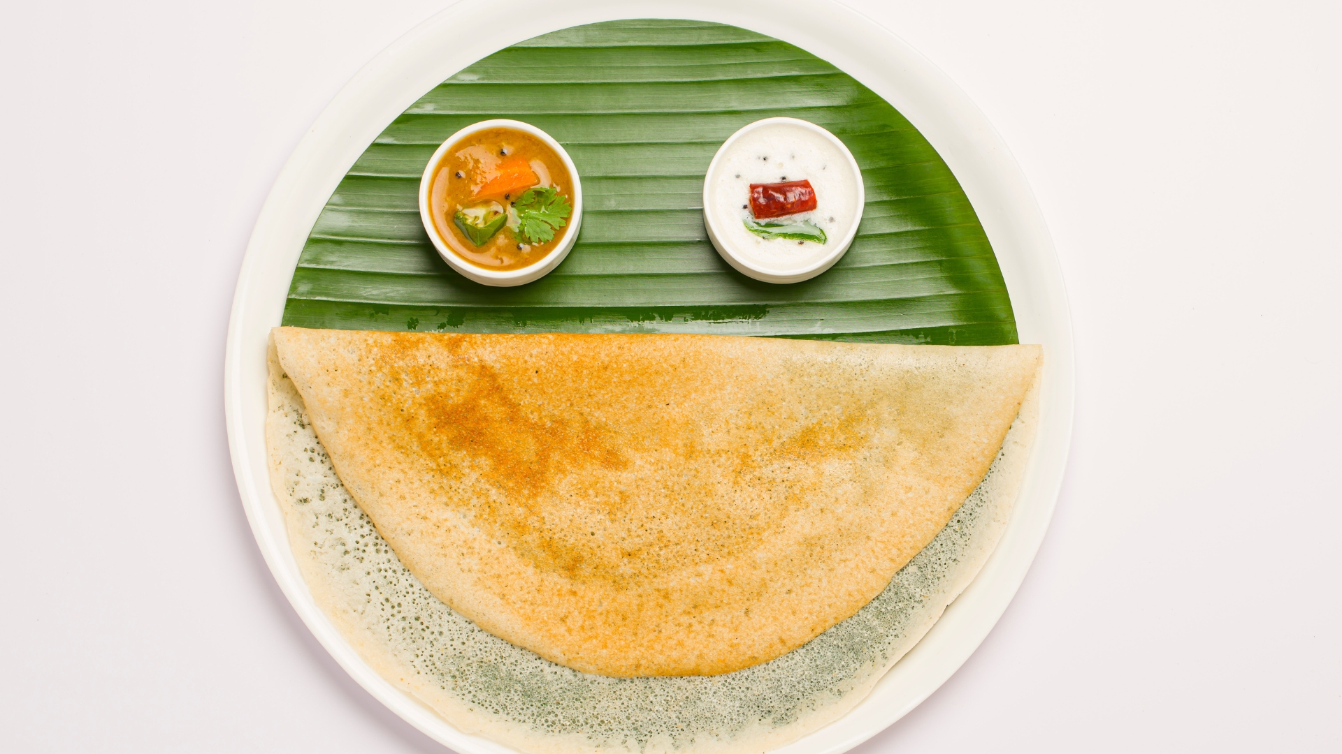 A golden-brown dosa served on a banana leaf with two small bowls of sambar and coconut chutney, arranged to resemble a smiling face.