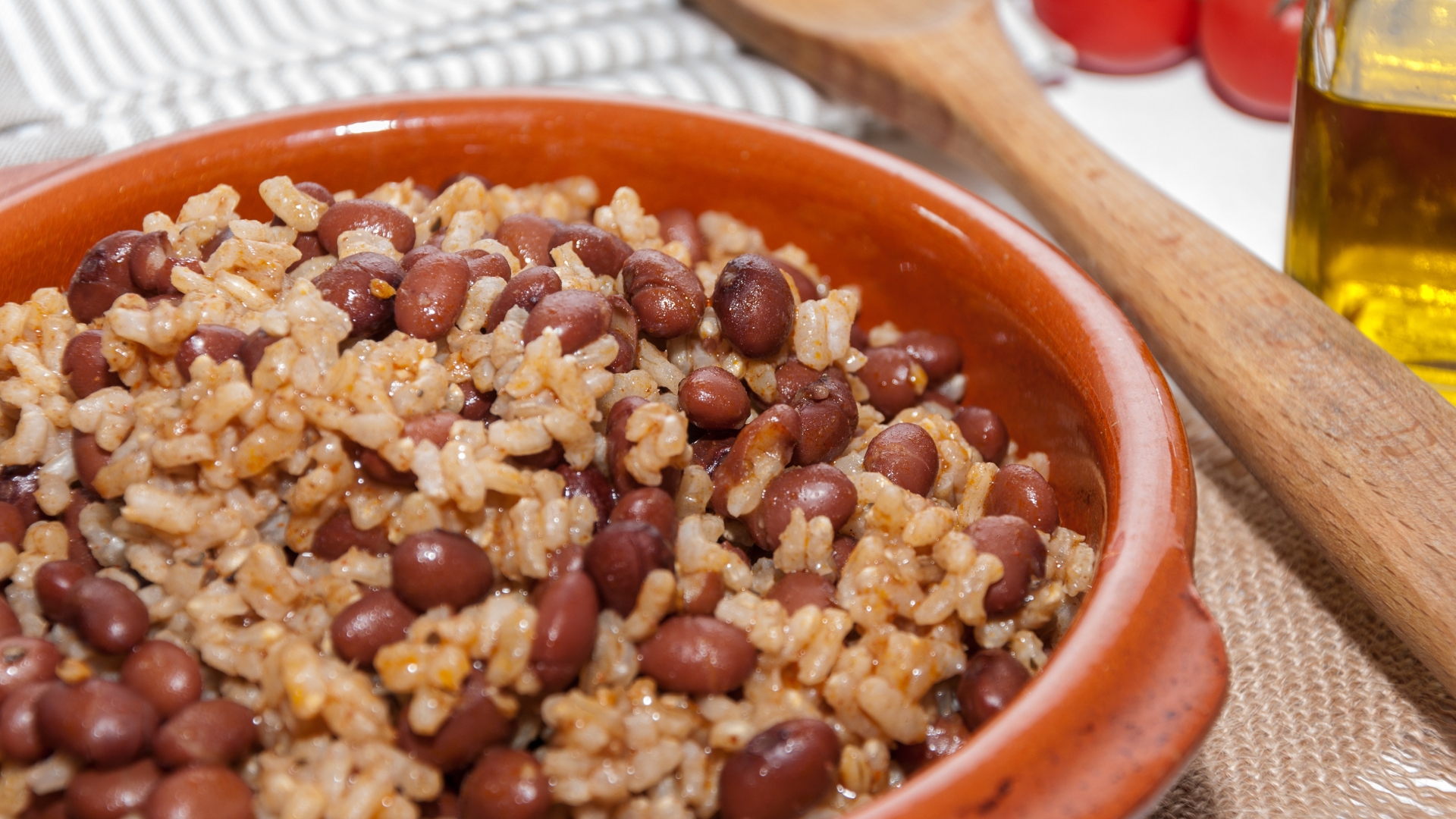 A bowl of red beans and rice, garnished with spices, next to a wooden spoon and fresh tomatoes.