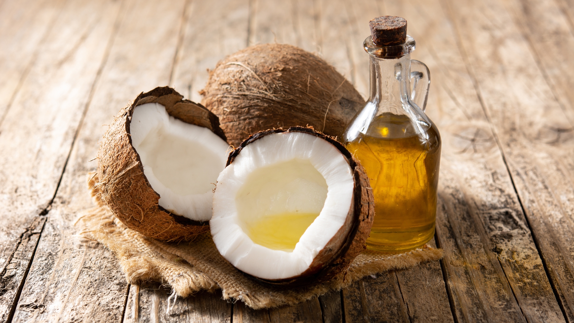 A glass bottle of coconut oil next to halved coconuts on a rustic wooden table.