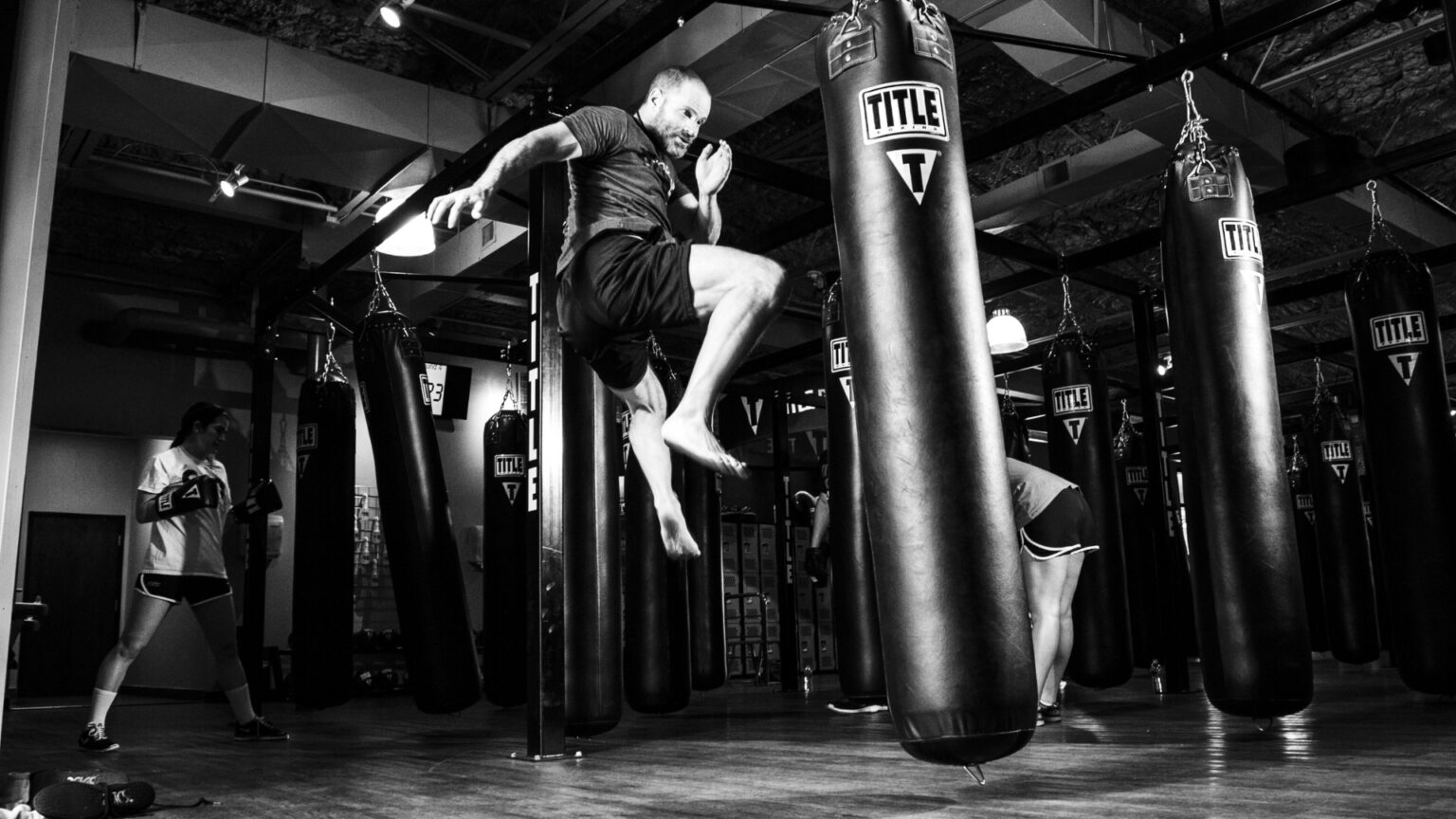 A fighter performing an explosive knee strike to a heavy bag in a boxing gym, demonstrating anaerobic power and agility.