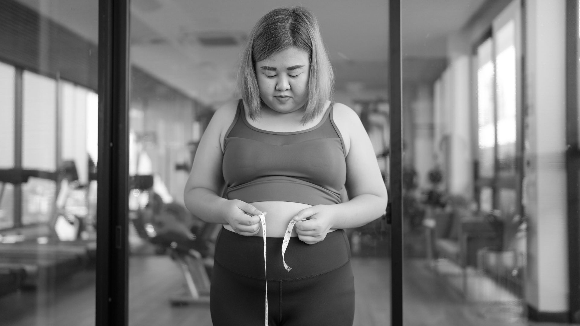 A woman in athletic wear standing in front of a gym mirror, looking down at a measuring tape around her waist, reflecting on her progress and weight loss journey.