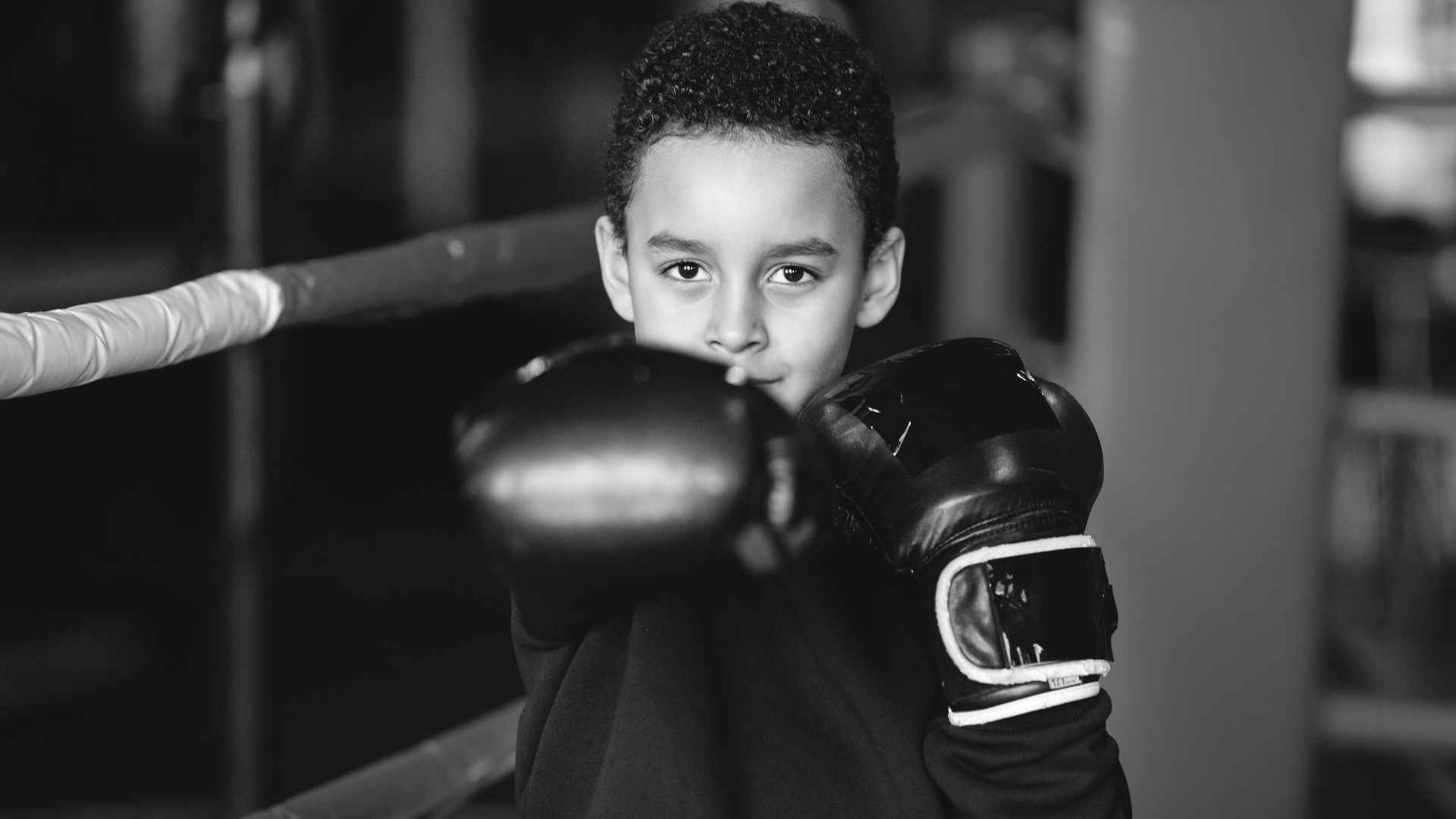 A young boy wearing boxing gloves in a gym, confidently posing with his fists up.